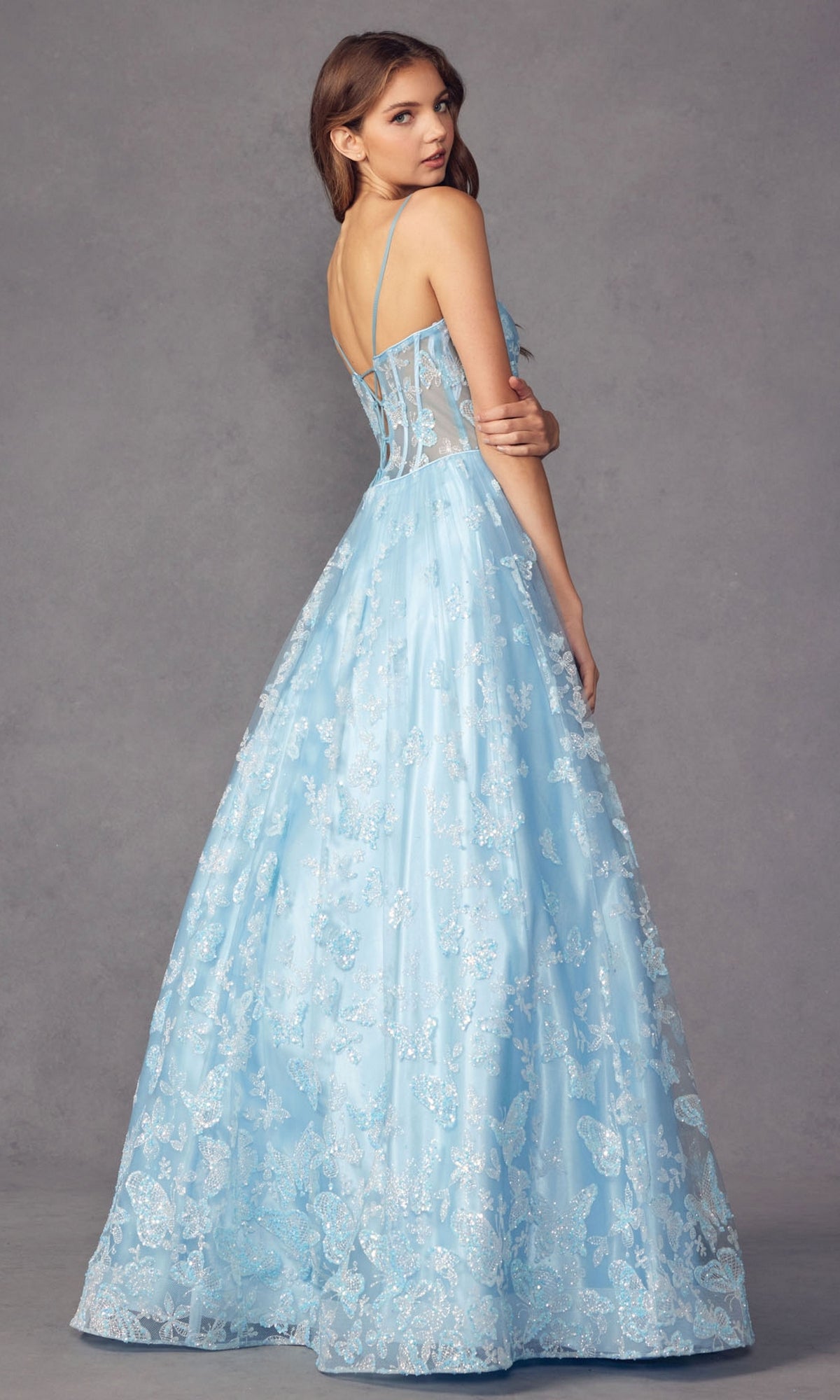 A woman stands with her back turned, wearing the Sheer-Corset Glitter-Print Long Prom Dress 2413, a light blue, floor-length A-line gown with sleeveless lace and floral details against a plain gray background.